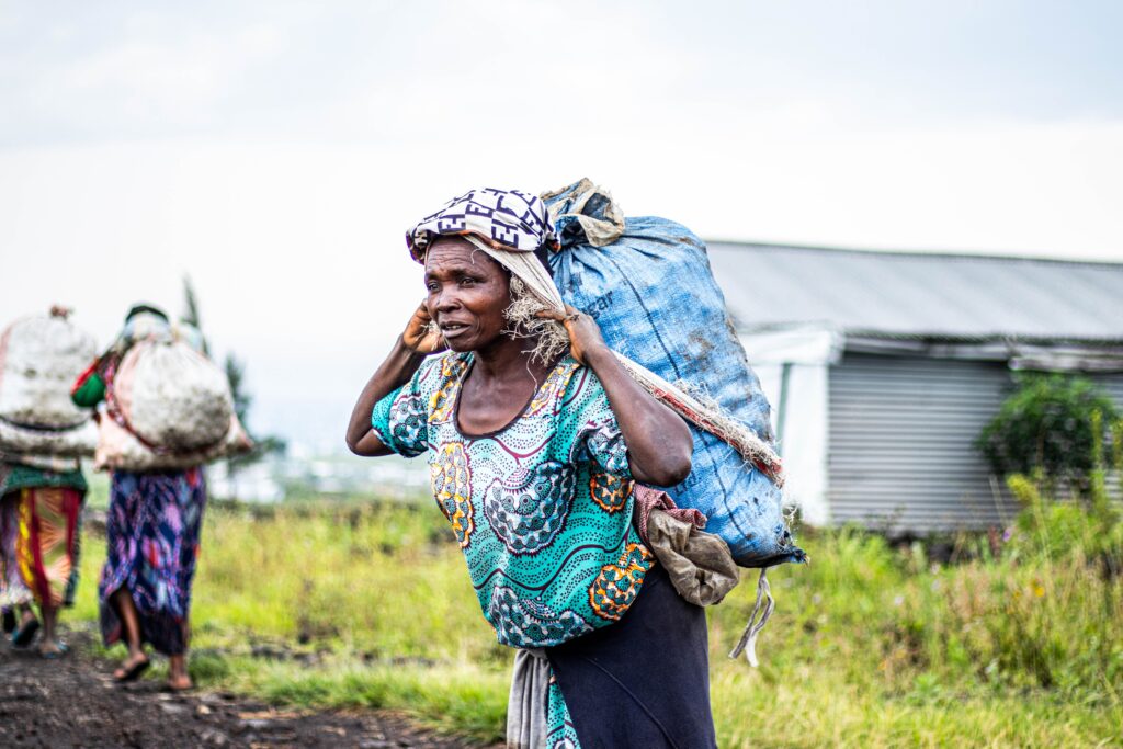 African women walking outdoors carrying heavy loads. Vibrant attire and rural setting.